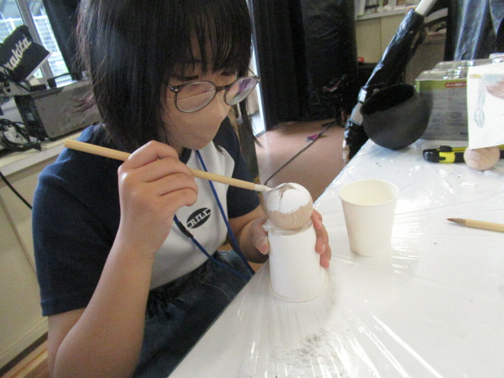 作業室内で眼鏡をかけた塾生が、紙コップの上に置かれた球体状の木製パーツに、細い筆を使って白い塗料を一点一点丁寧に塗り込んでいる写真