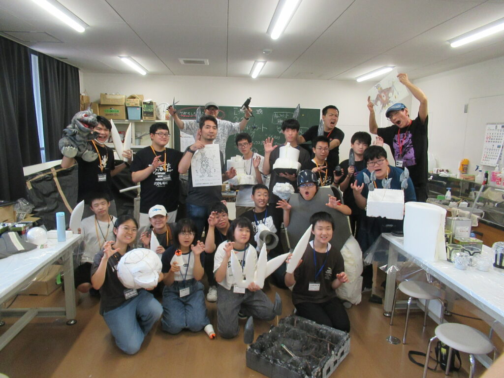 作業室内で、完成した怪獣の頭部や鋭い爪のパーツを手に持った塾生と講師陣が、怪獣になりきったポーズや笑顔でカメラを囲んでいる集合写真