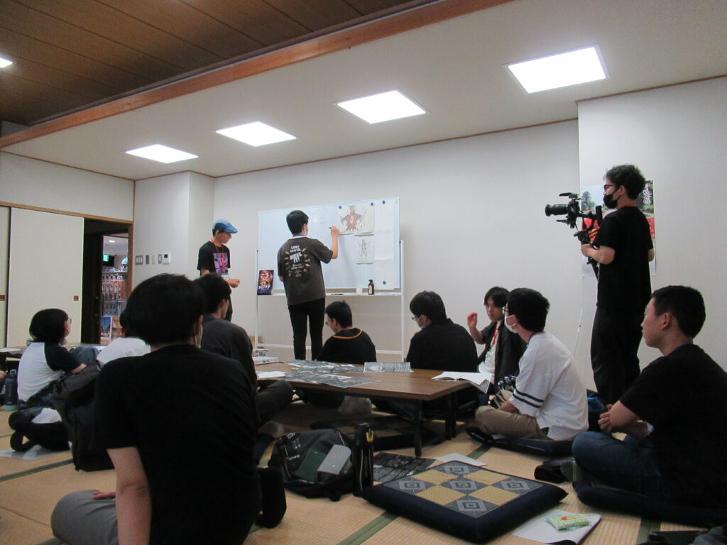 和室の研修会場で、数人の若者たちがホワイトボードに貼られた怪獣のイラストを囲んで議論し、その様子を後方から座卓に座った塾生やビデオカメラを構えた撮影スタッフが見守っている写真