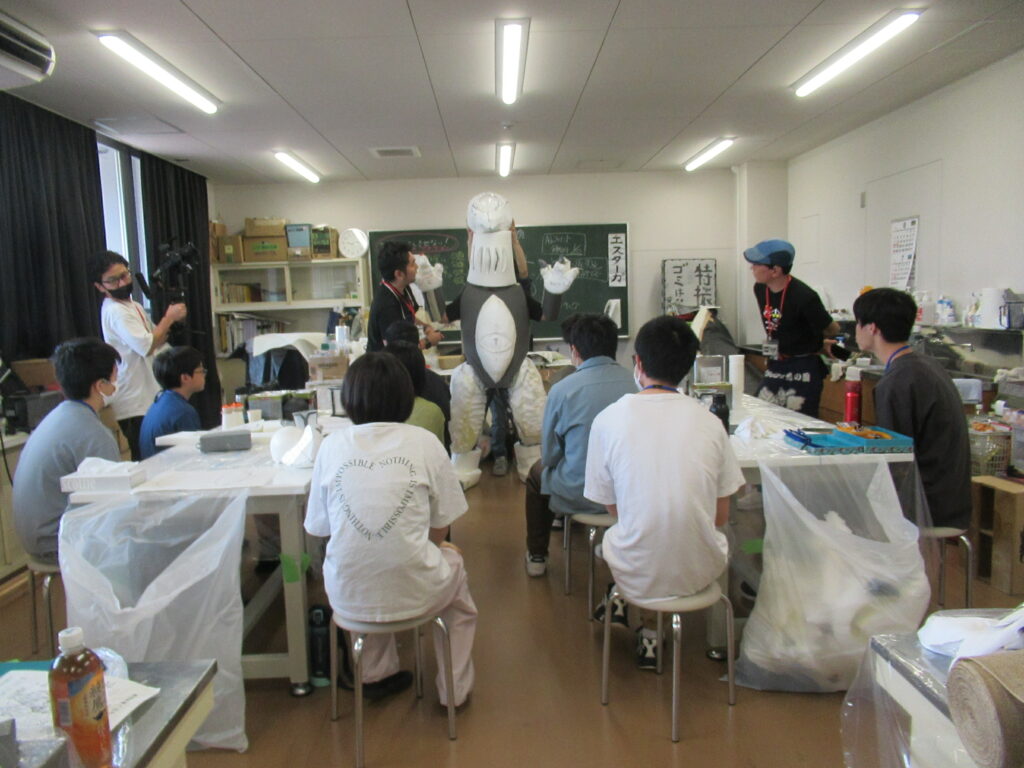 作業室内で、塾生が作業机を囲んで座っており、中央で講師が未塗装の白い怪獣の着ぐるみ組み立てて、解説している風景を捉えた写真