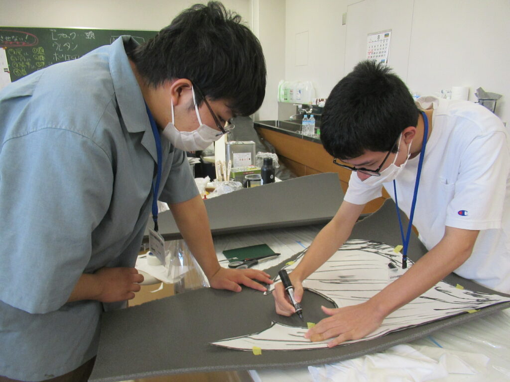 作業室内で、ウレタンスポンジを手で押さえている男性と、白Tシャツを着た男性が灰色のウレタンスポンジの上に置かれた型紙の縁に沿って、黒いマーカーで丁寧に下書き線を書き写している写真