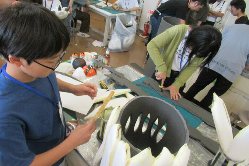 作業室内で、男の子が製作中の造形物に刷毛で接着剤を塗り、傍らで緑のカーディガンを着た女の子はカッターを使って灰色のウレタン素材を慎重に切り抜いている写真