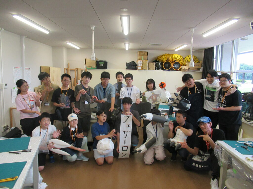 作業室内で、「フェザーロン」と書かれた紙を持つ中央の男性を囲み、制作した怪獣の爪や牙などのパーツを手にしたり、怪獣を模したポーズを取ったりしている集合写真