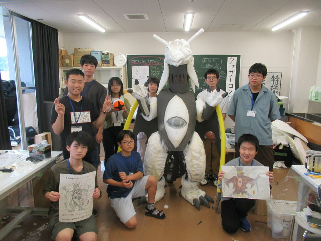 屋内の教室で、中央に立つ白と黒の大きな怪獣の着ぐるみを囲むように、設計図やイラストを手に持った塾生が集まって笑顔でポーズを取る集合写真