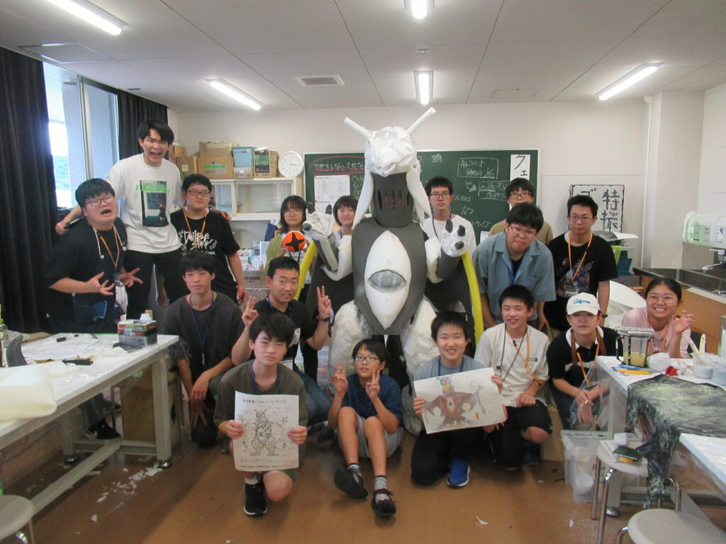 屋内の教室で、白とグレーを基調とした大きな怪獣の着ぐるみを中央に配置し、その周囲で設計図やイラストを掲げた塾生が集まった集合写真