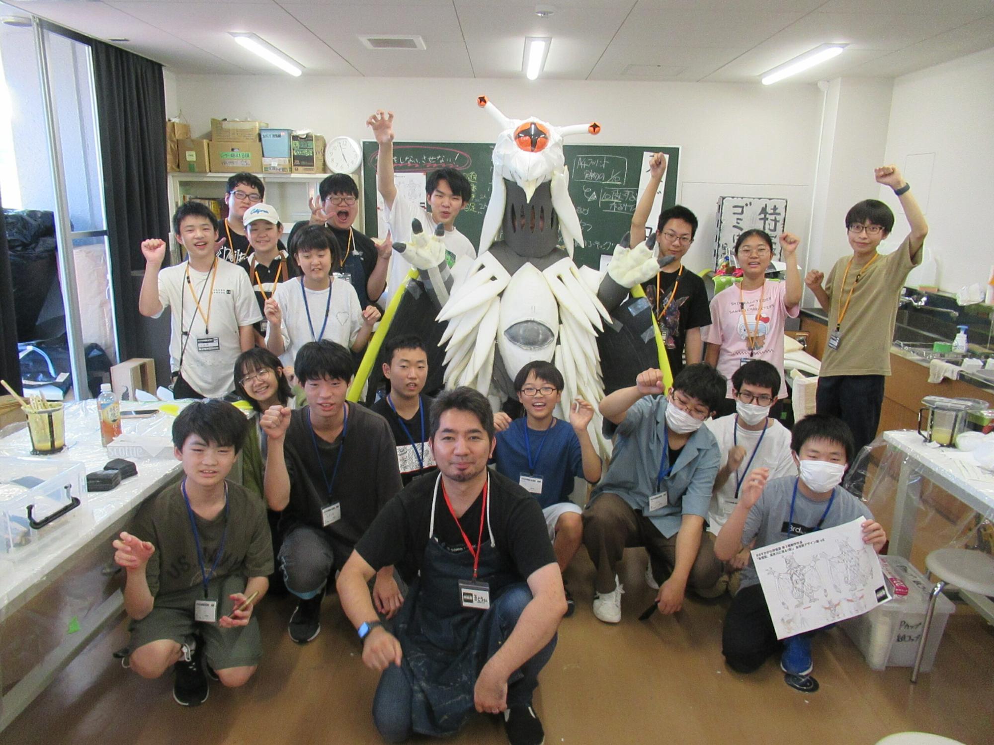 作業室内で、中央にそびえ立つ白と黒の巨大なオリジナル怪獣の着ぐるみを囲み、制作に携わった多くの塾生と講師陣が、拳を掲げたり笑顔を見せている集合写真