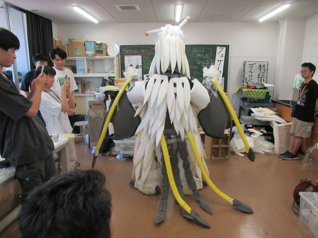 屋内の教室で、白とグレーのパーツや黄色い触角のような長い突起を持つ怪獣の着ぐるみを背後から捉え、その周囲で数人の塾生がスマートフォンを向けて撮影したり、仕上がりを確認したりしている制作風景の写真