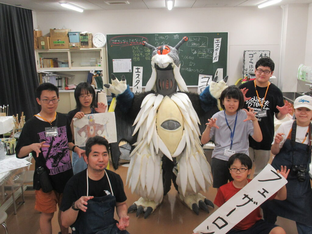 屋内の教室で、中央に立つ白とグレーの大きな怪獣の着ぐるみを囲むように、設計図やイラストを手に持った塾生が集まって笑顔や真剣な表情でポーズを決めている集合写真