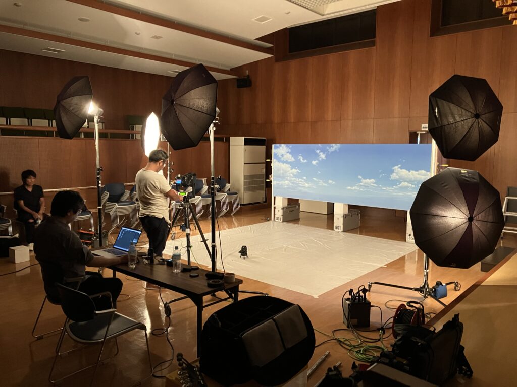 木目調の壁に囲まれた広い室内で、青空と白い雲が描かれた横長の背景パネルを設置し、複数のアンブレラ型照明やカメラ、PCを操作するスタッフを配置して特撮作品の撮影現場の様子を捉えた写真