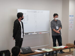 ホワイトボードの前に男性二人が立ち、手前の机にはノートパソコンや資料が並べられている写真