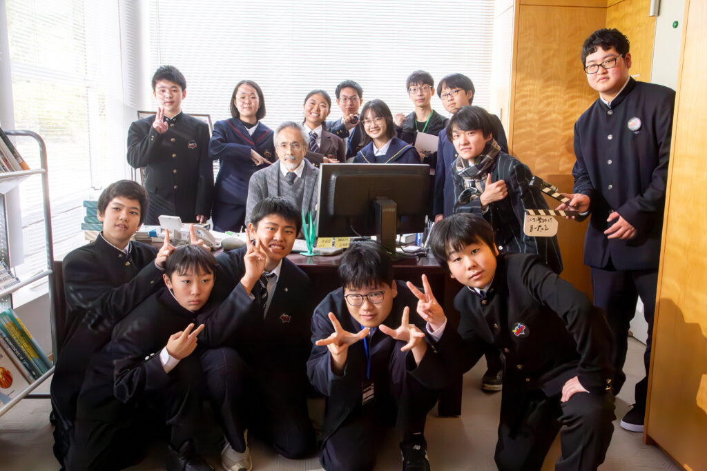 塾生たちが役者の男性が座った机を囲んで集まり、ポーズをとっている記念写真