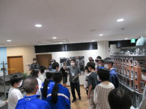 展示室内に置かれた精巧な建物模型や資料の横で、講師が多くの塾生たちに身振り手振りを交えながら解説を行っており、塾生が熱心に耳を傾けている写真