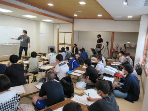 畳敷きの和室で多くの塾生が机に向かって座り、前方のホワイトボードを使って解説する講師の話を熱心に聞きながらメモを取っている写真
