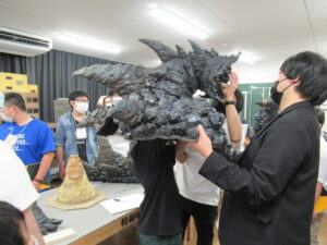 岩肌のような質感を持つ黒く巨大な怪獣の頭部や背びれの造形物を塾生に被せている写真