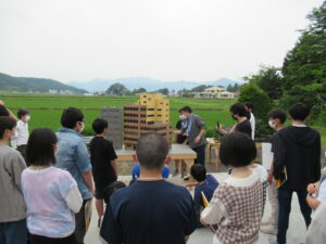 田園風景を背景にした屋外で、机の上に置かれたビルなどの精密なミニチュア模型を囲みながら、講師の解説を熱心に聞き入っている塾生の写真