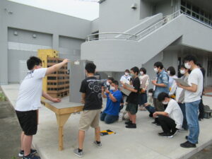 屋外のコンクリート敷きのスペースで、テーブルの上に設置されたビル群のミニチュア模型を調整する塾生と、その様子を見守っている写真