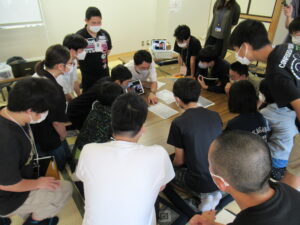 和室で、多くの塾生たちが机を囲んで座り、机の上の資料をのぞき込み、中央にいる男性の話を聞き入っている様子の写真