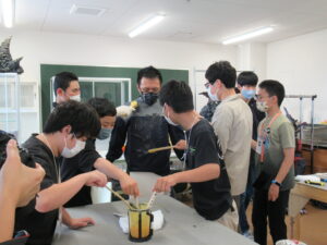作業室で、数人の塾生たちが容器に入った液体を棒で混ぜたり、怪獣の着ぐるみを着用した男性に黒色の液体を塗布している写真