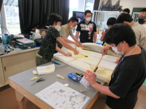 作業室で、数人の塾生たちが机の上に広げた大きな白いウレタン素材に、刷毛を使って黄色い接着剤のような液体を丁寧に塗り広げている写真