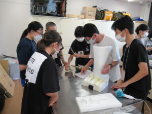 明るい作業室で、数人の塾生たちが机の上に置かれた白いウレタン素材を組み立てたり、中央の男性が指し示す図面を真剣に確認したりしながら、特撮用の造形物を制作している様子の写真
