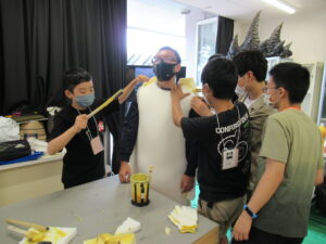 作業室で、数人の塾生たちが白いウレタン素材を男性の体に合わせながら、特撮用の着ぐるみのベースとなるパーツを丁寧にフィッティングしている様子の写真