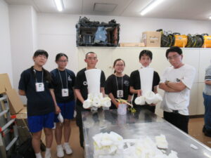 室内で、制作中の特撮用造形物を手に持った塾生たちと講師の男性が、横一列に並んで写した記念撮影の写真