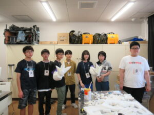 作業室で、制作した怪獣の鋭い爪がついた腕の造形パーツを手に装着して掲げ、横一列に並んで写した記念撮影の写真