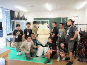 作業室で、白いウレタン素材を組み合わせて作られた怪獣の着ぐるみの胴体パーツを囲み、複数の塾生たちが集まって写した集合写真