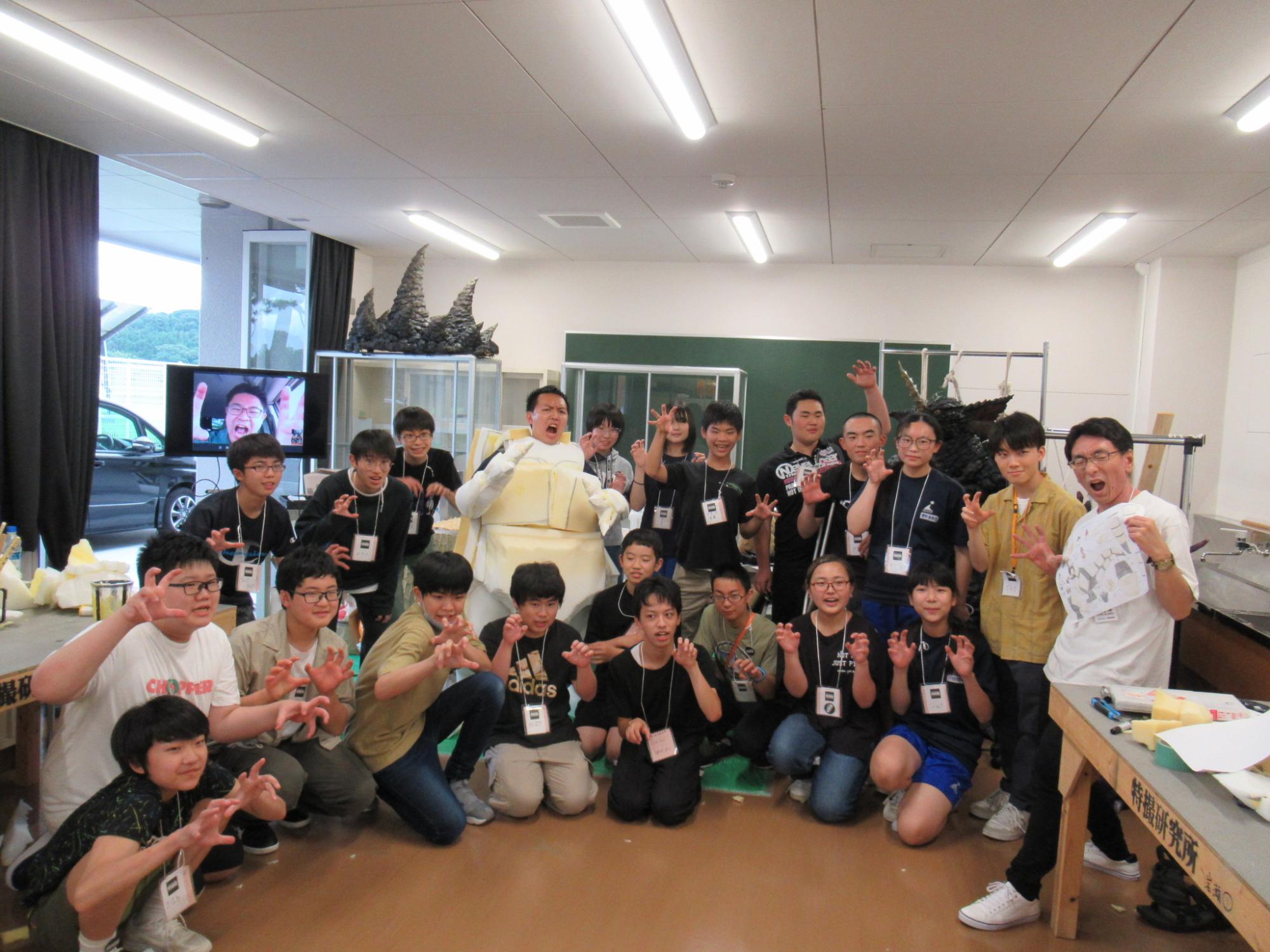 明るい室内で、怪獣の着ぐるみを囲むように集まった大勢の講師や塾生たちが、カメラに向かって怪獣のポーズをしながら写した集合写真