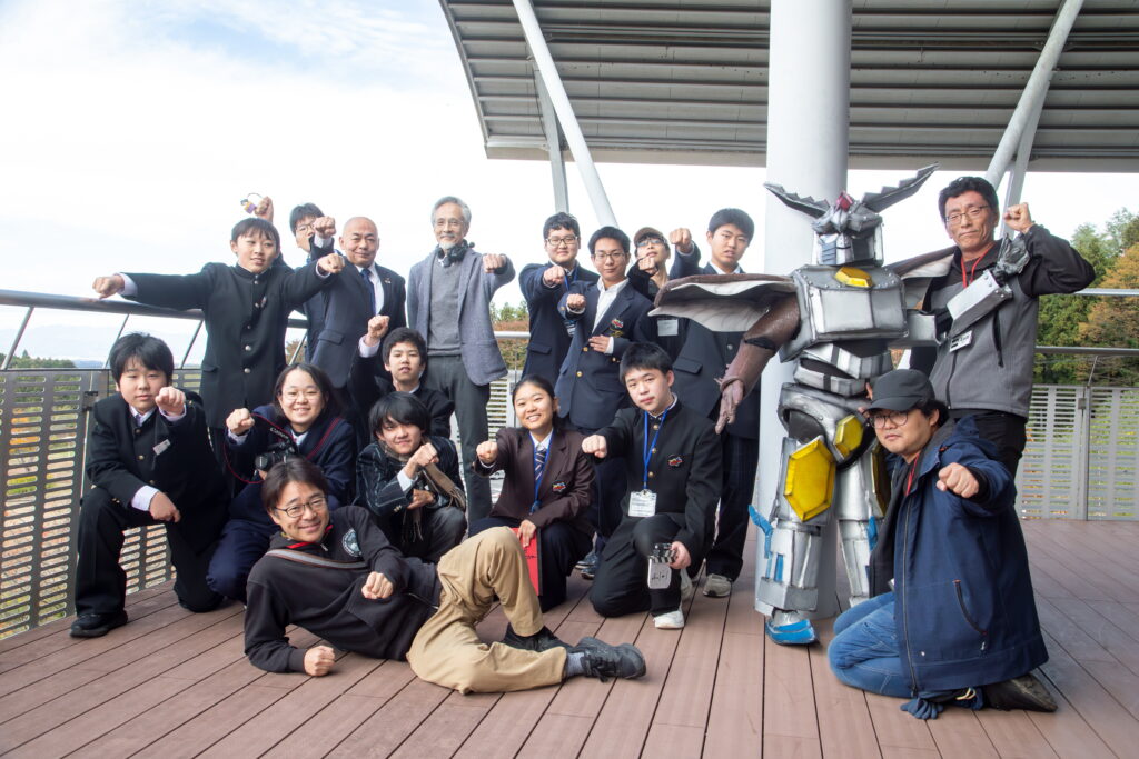 多数の参加者がロボット風の大型キャラクターと並び拳を突き出して力強いポーズを決めている集合写真