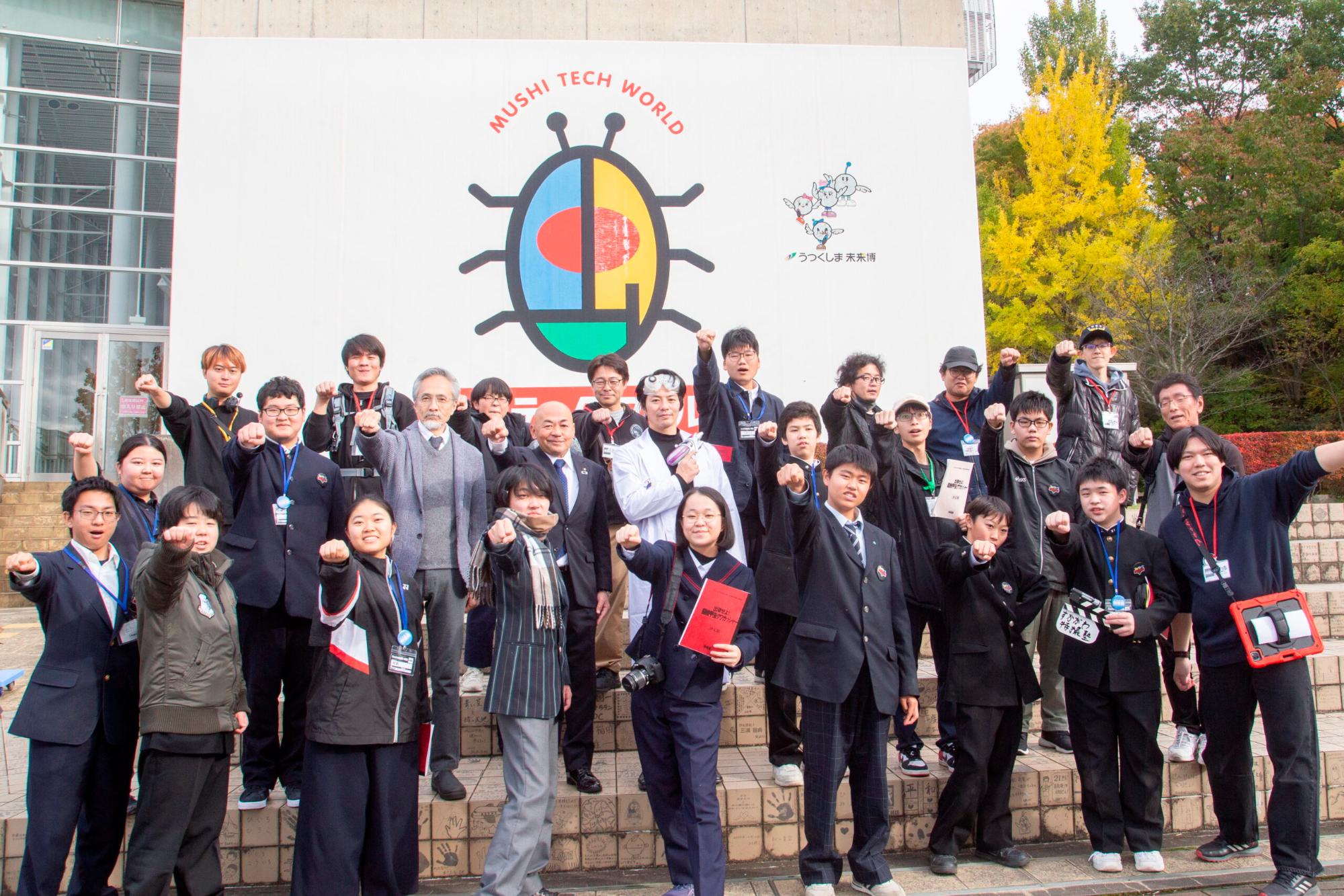 建物前に集まった多数人が名札を着け拳を掲げて整列し、背後にMUSHI TECH WORLDのロゴ看板が見える集合記念の様子を撮影した写真