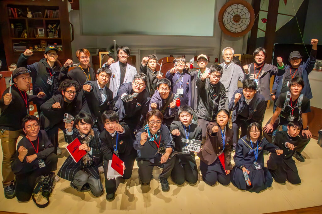 多数の参加者が撮影セットに集まり拳を力強く突き上げてポーズを決めた集合写真