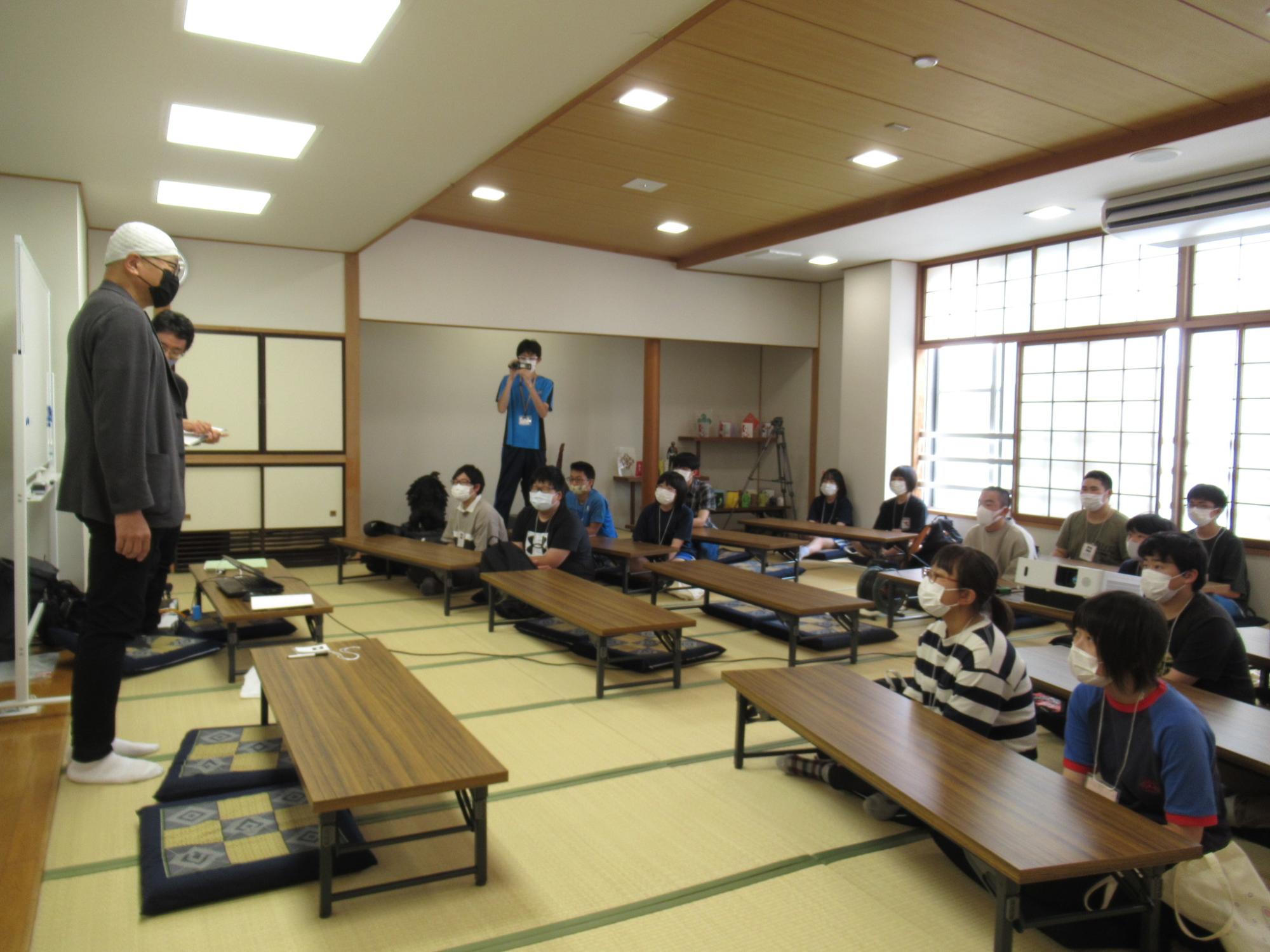 畳敷きの和室で、マスクを着用した大勢の塾生が長机の前に座り、前方でホワイトボードを背に話をする講師の男性の説明を真剣に聞いている写真