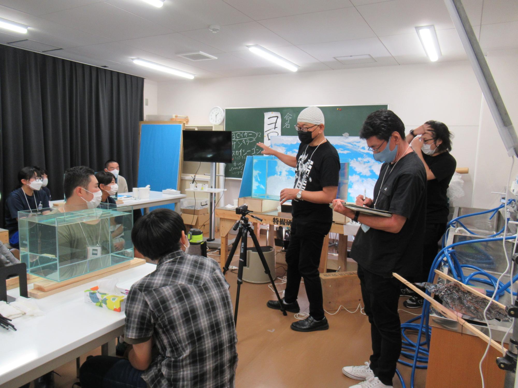 屋内の教室で、水槽が置かれた机の前に座る塾生に対し、黒いTシャツに白いニット帽を被った講師の男性が、背後の黒板や青空の背景板を指差しながら解説を行い、傍らで別のスタッフがメモを取っている講習風景の写真