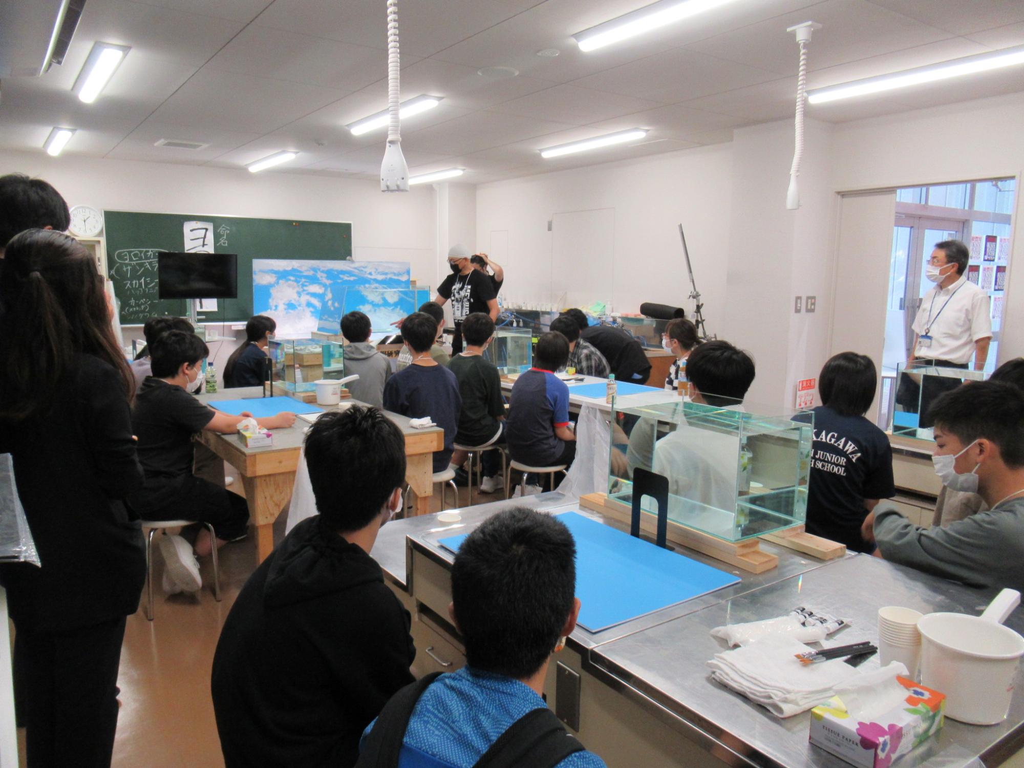 明るい教室の中で、机に置かれた複数の小さな水槽を囲むように大勢の塾生が座り、前方で雲の背景板を背に解説を行う講師の男性の話を真剣な表情で聞いている実習風景の写真