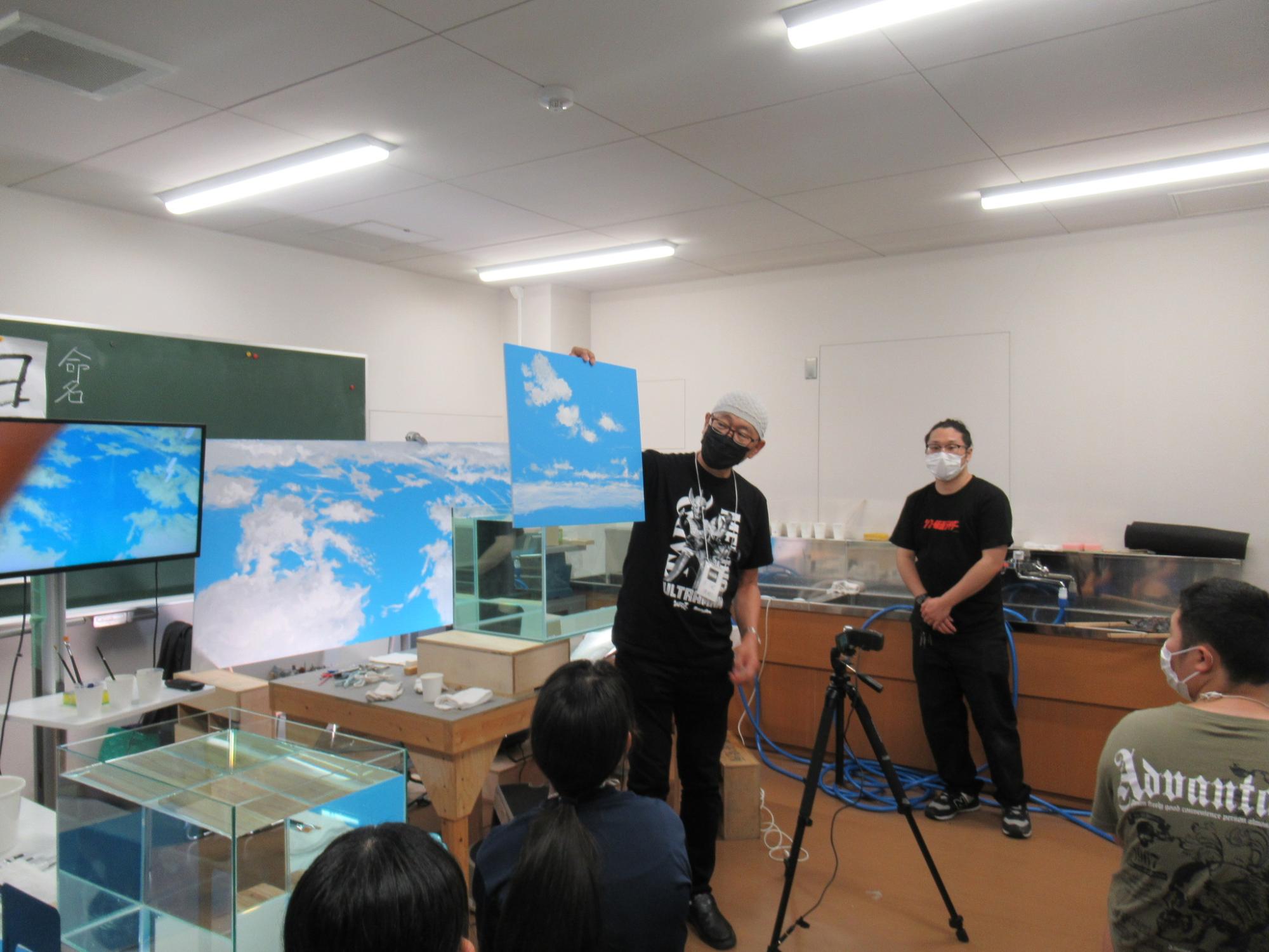 屋内の教室で、黒いTシャツに白いニット帽、黒いマスクを着用した講師の男性が、青空と白い雲が描かれた背景板を塾生の前で高く掲げ、参加者達に説明を行っている写真