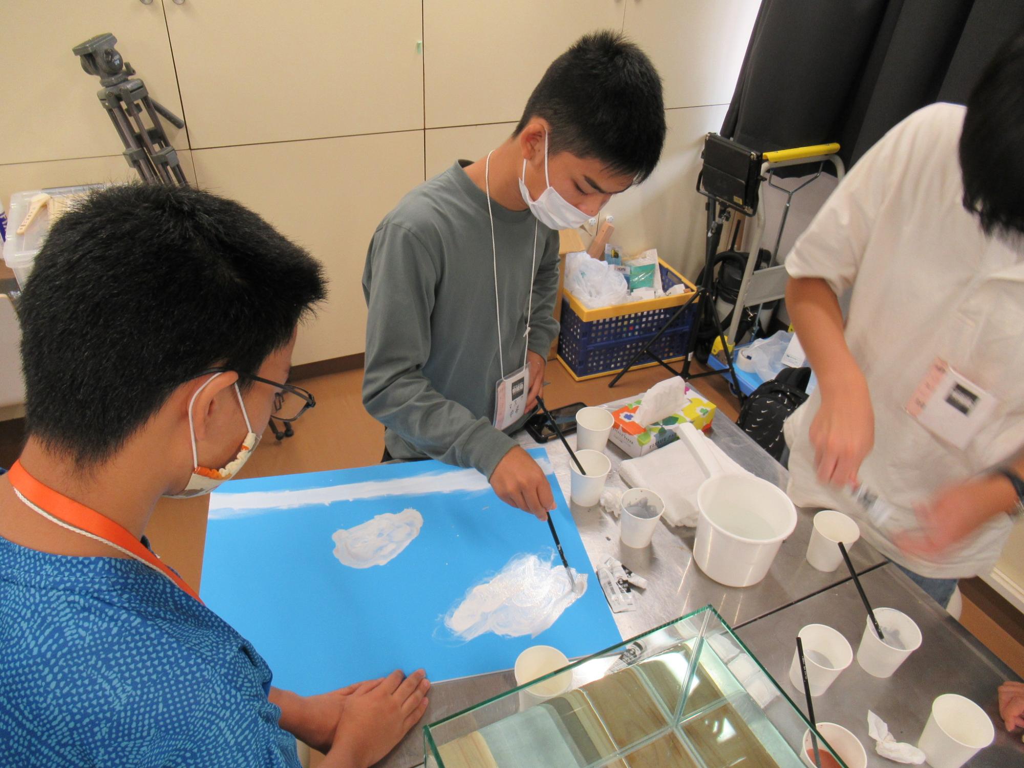 明るい教室の中で、机に置かれた水色のパネルに筆を使って白い雲を描き込む塾生の手元を捉えた写真