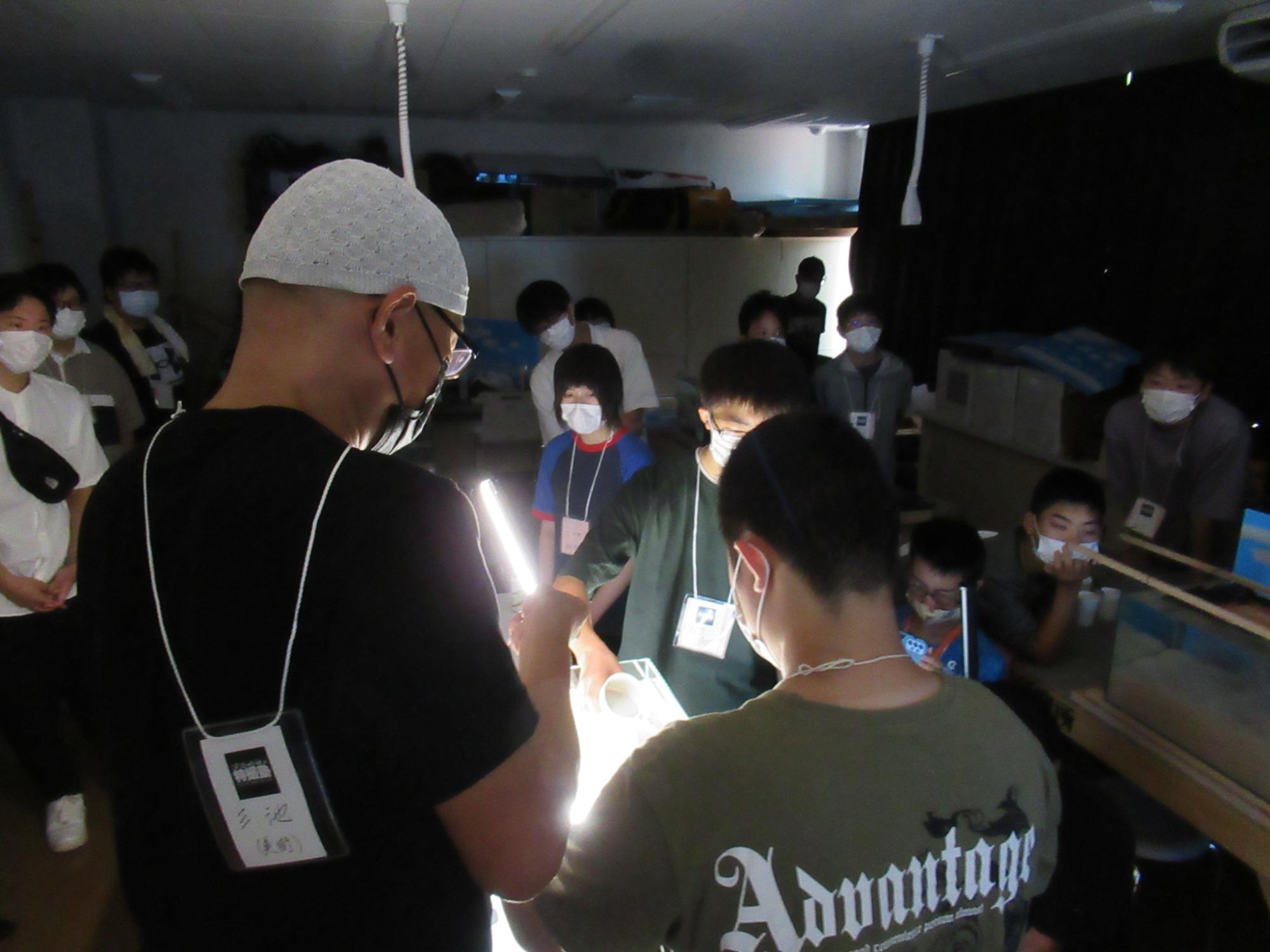 薄暗い教室の中で、白いニット帽を被った講師の男性がLEDライトを手に持ち、水槽が置かれた机を照らしながら説明を行っている写真