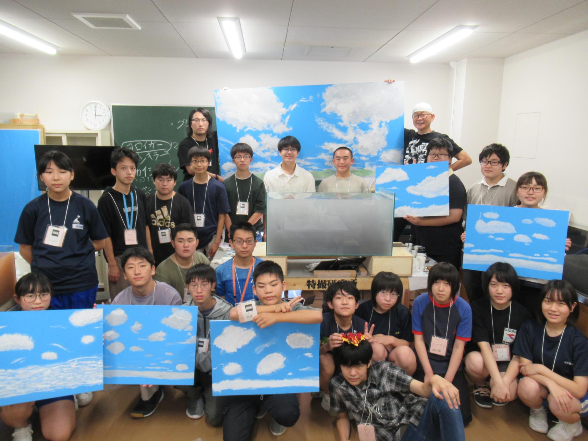屋内の教室で、青空と白い雲が描かれた大小さまざまな背景板やイラストを手に持った大勢の塾生が、中央の水槽を囲んで並んでいる集合写真
