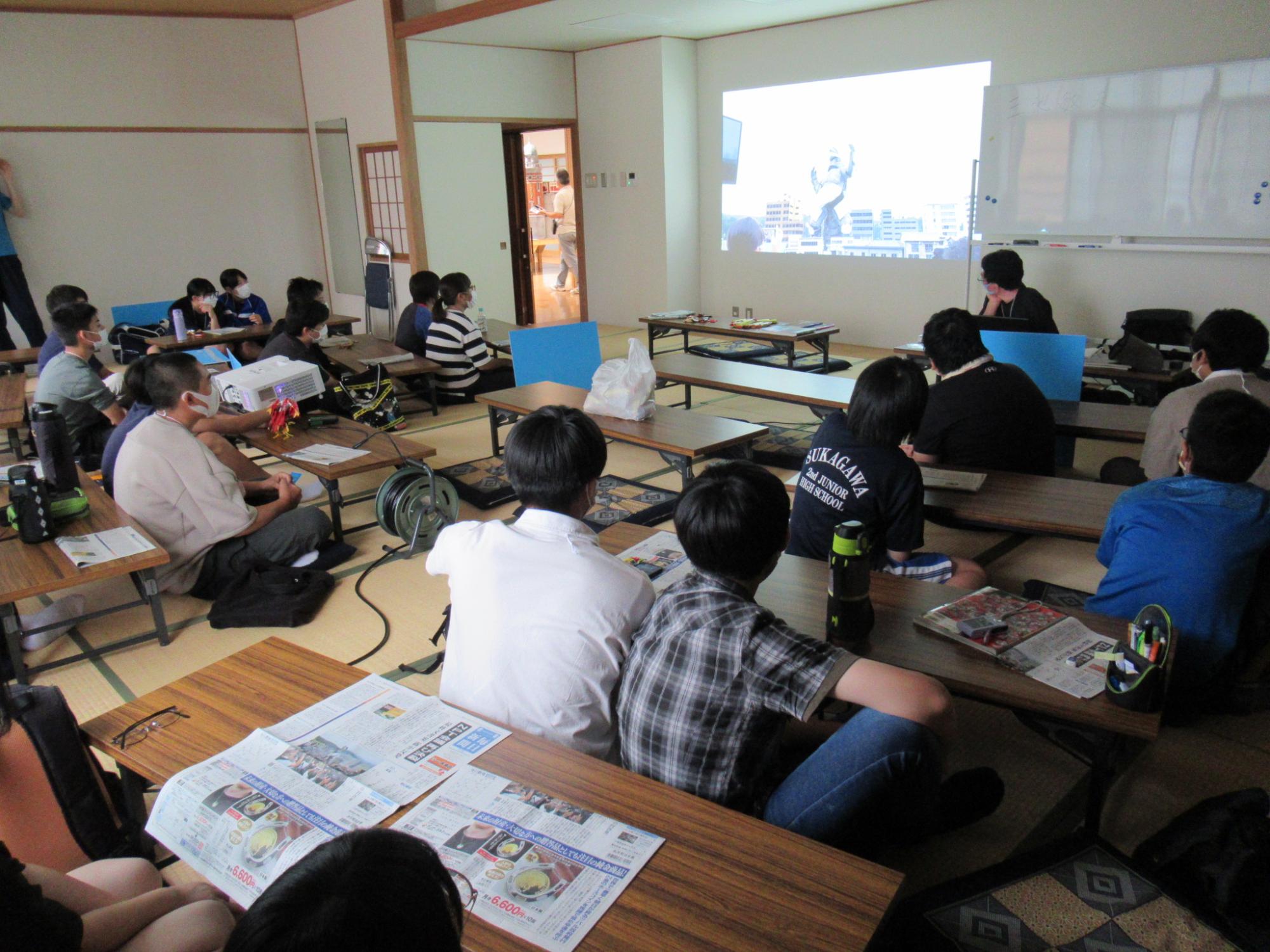 畳敷きの和室で、長机の前に座った多くの塾生が、壁に投影された特撮映像のプロジェクター画面を真剣な表情で見ている写真