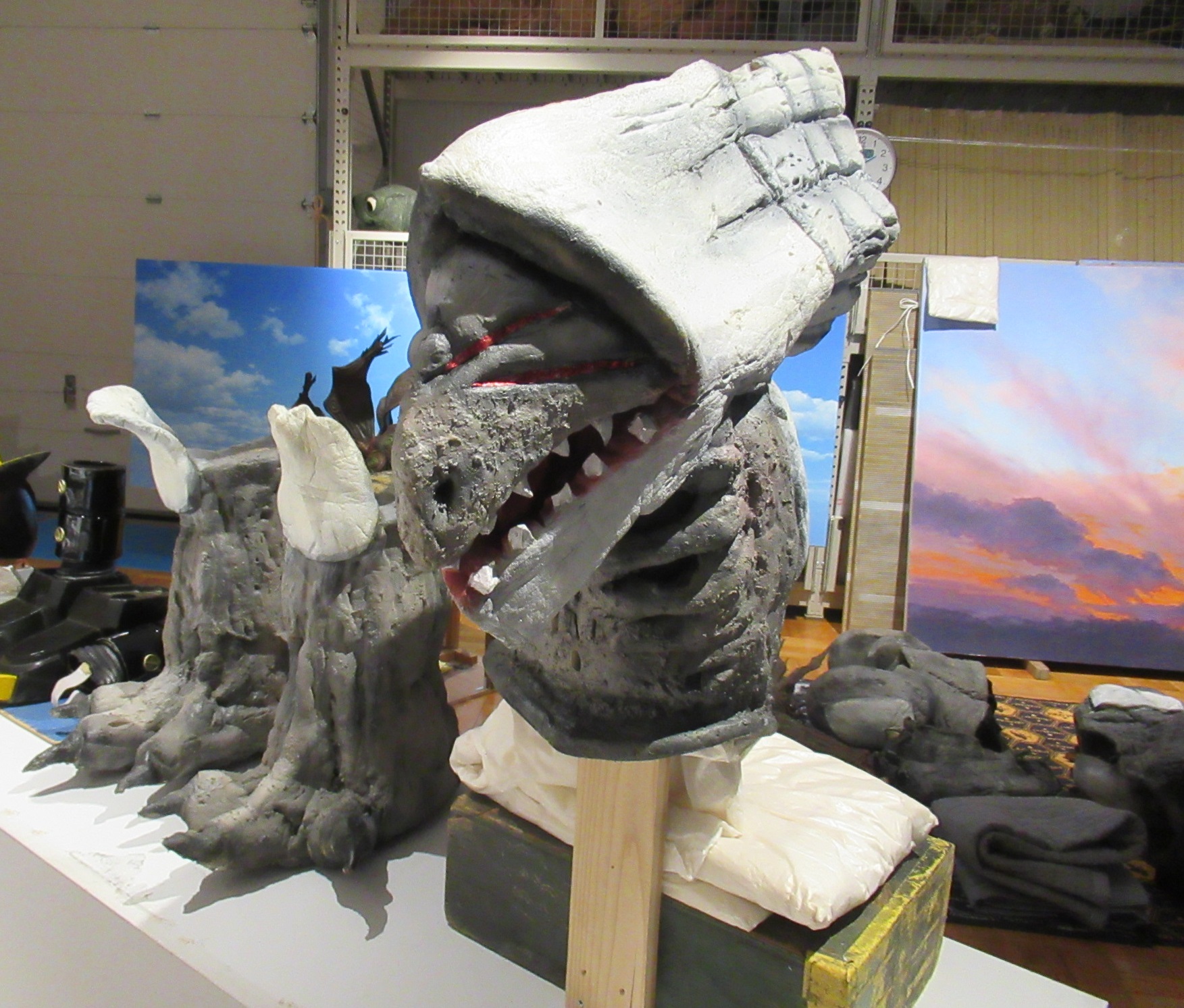 作業場の台上に灰色の怪獣風頭部模型が置かれ周囲に手足のパーツや背景画が並ぶ写真