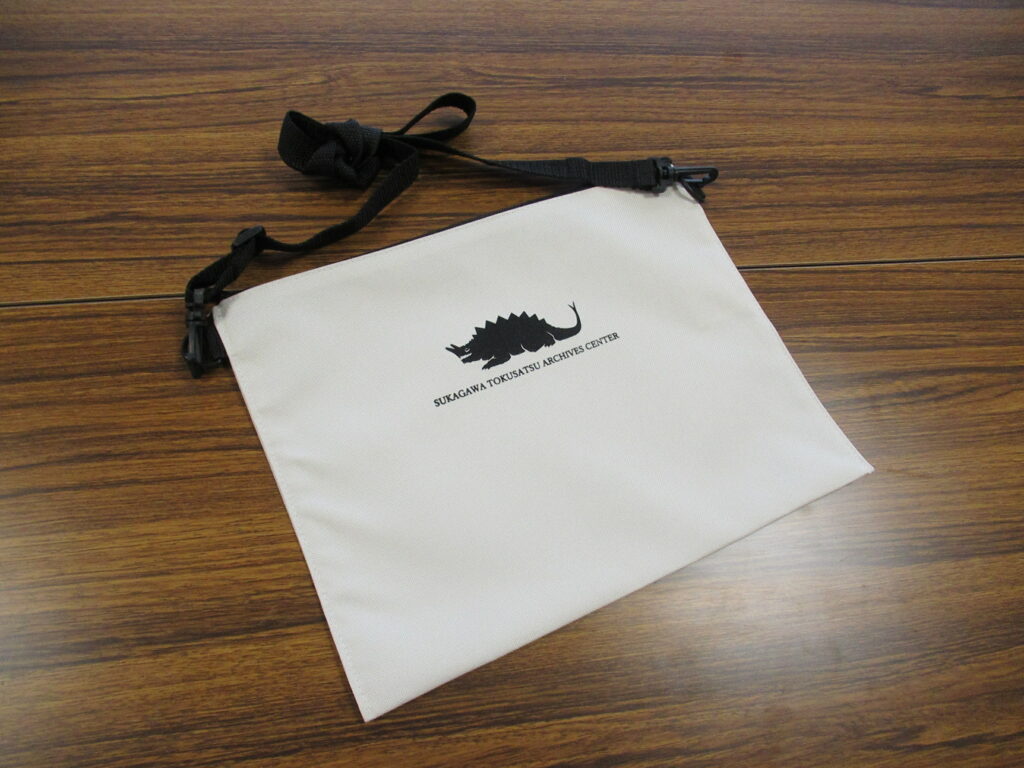 机の上に、背中に鋭い突起を持ち、頭部から角のようなものが生えた巨大な怪獣のシルエットと「SUKAGAWA TOKUSATSU ARCHIVES CENTER」の文字が印刷された白地のサコッシュの写真