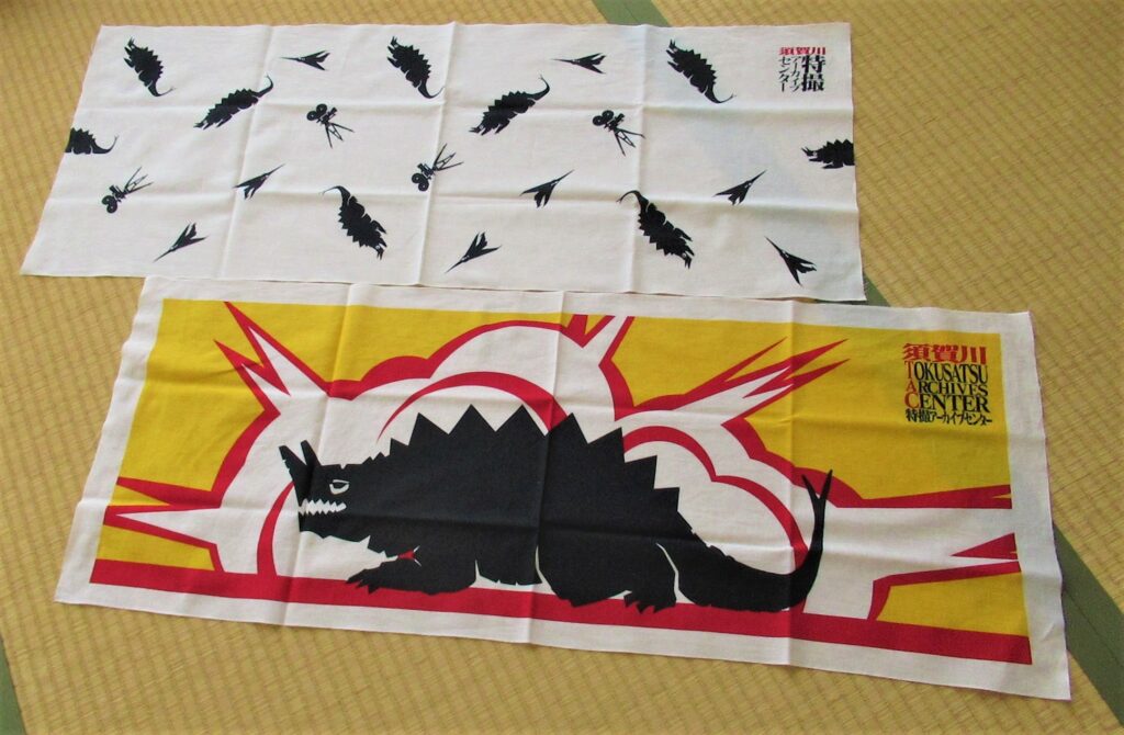 畳の部屋に並べて広げられた二種類の手ぬぐいがあり、上には怪獣やカメラの小さなシルエットが散りばめられた白いもの、下には 背中に鋭い突起を持ち、頭部から角のようなものが生えた巨大な怪獣のシルエットが描かれた黄色と赤の鮮やかなオリジナル手ぬぐいの写真