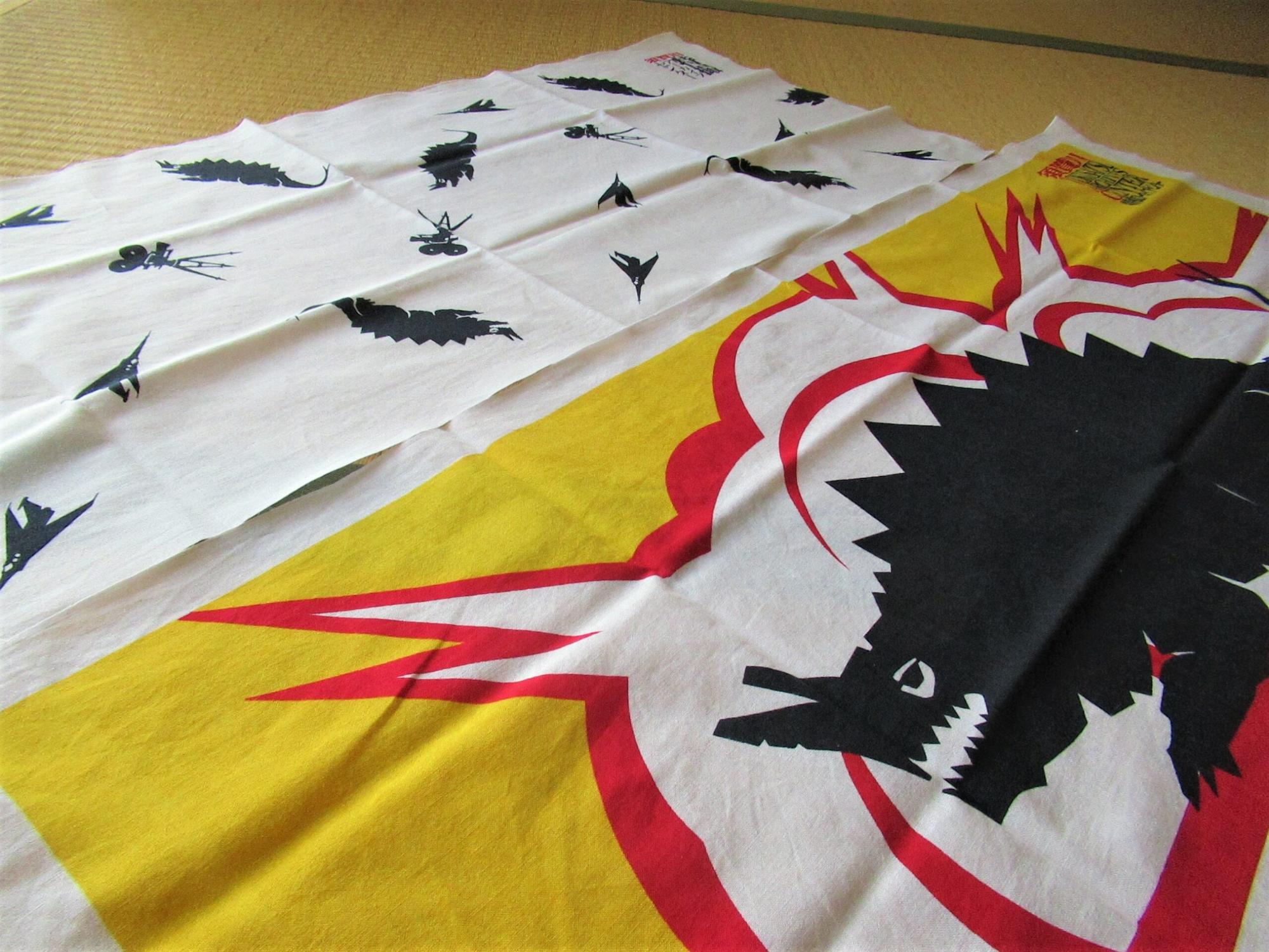 畳の部屋に、背中に鋭い突起を持ち、頭部から角のようなものが生えた巨大な怪獣のシルエットと赤い爆発のようなグラフィックが大きく描かれた黄色の手ぬぐいと、小さな怪獣やカメラのアイコンが等間隔に並んだ白い手ぬぐいの二種類が広げて置かれている写真