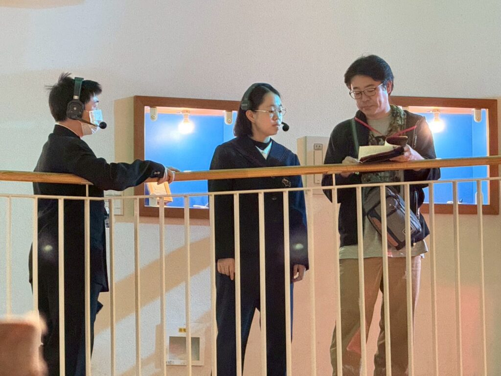 手すりの前でインカムを着けた二人が並んで立ち、講師の男性が手元の資料を示して会話をしている様子の写真