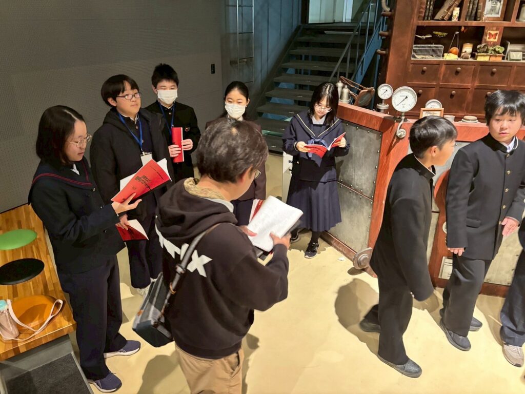 複数の塾生が赤い冊子を手にセットの前に立ち、指導者が資料を確認しながら説明を進めている様子の写真