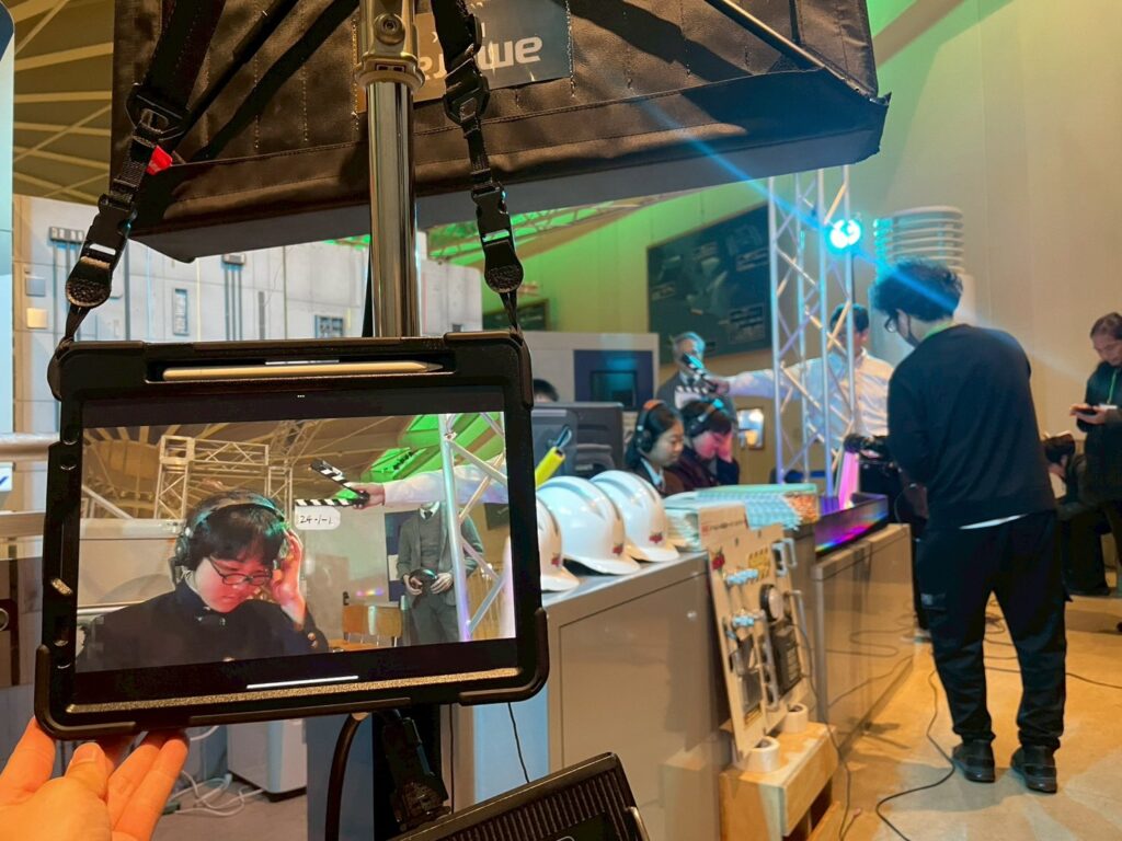 手前のタブレット画面にインカムを着けた塾生の映像が映し出され、その奥のセットでカメラの撮影が行われている様子の写真