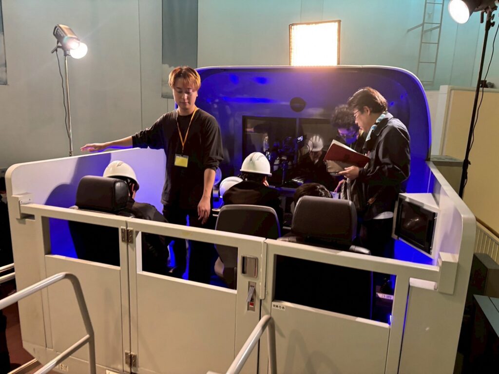 ヘルメットを着用した塾生たちが操縦席風のセット内で装置の操作手順を確認し講師らが側面から様子を見守りつつ資料を確認している様子の写真