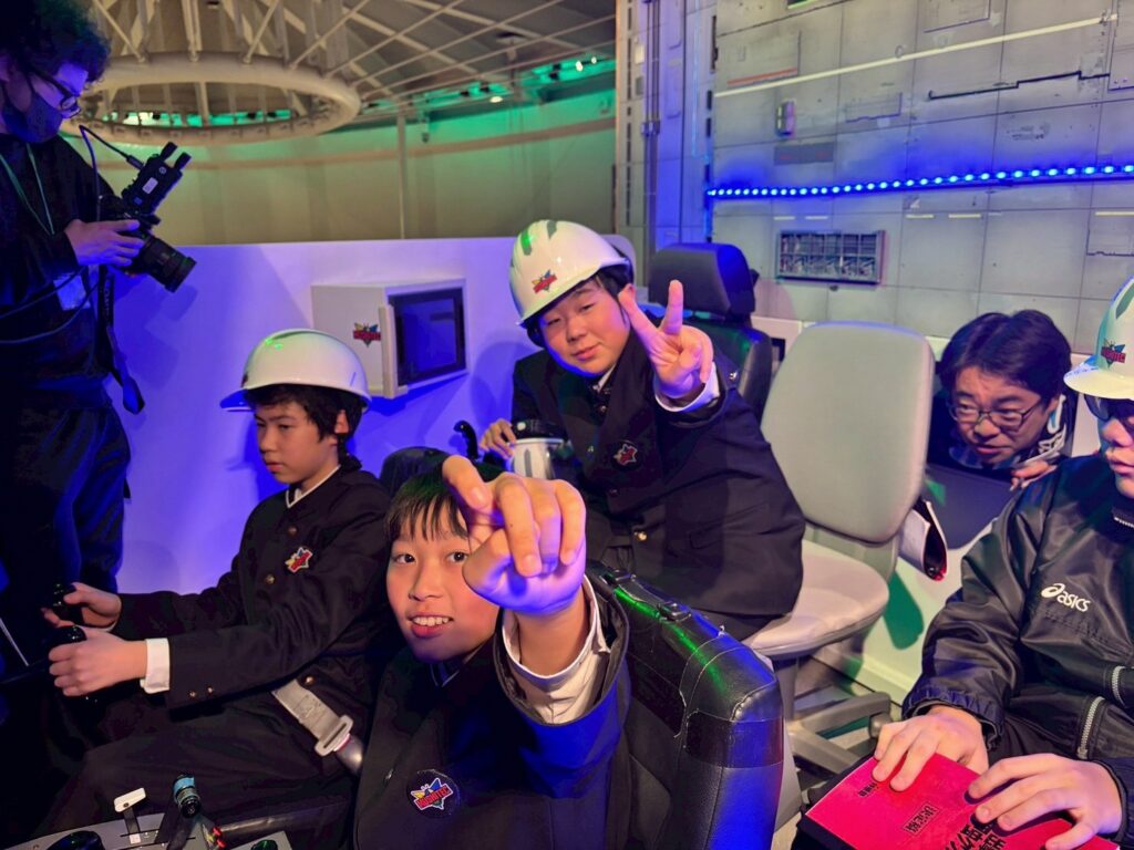 ヘルメットを着用した数人の塾生が操縦席風のセットに座り、カメラに向かってポーズをとっている写真