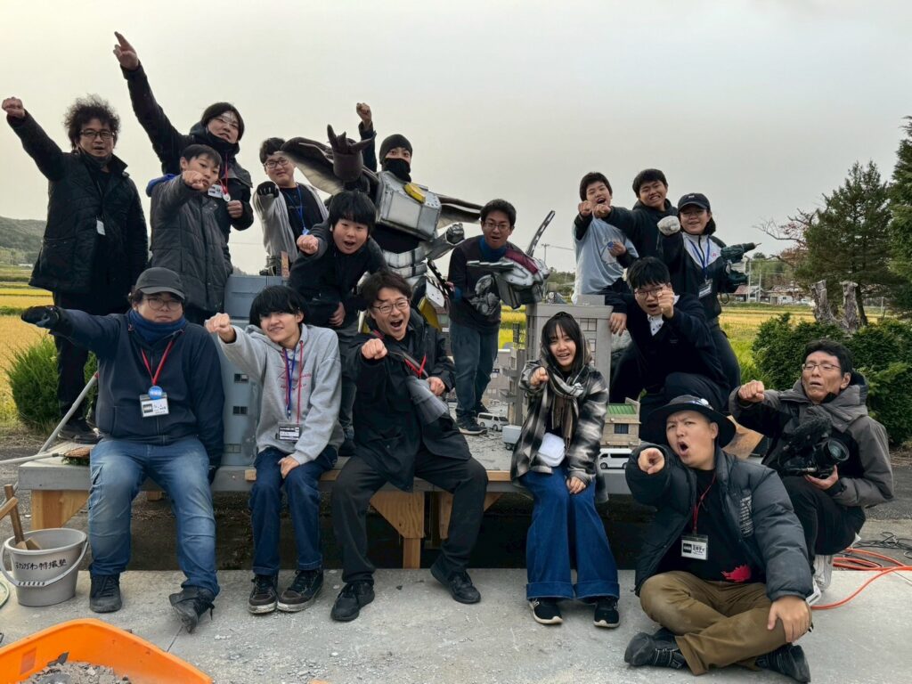 屋外で、多数の塾生やスタッフがロボット風スーツの演者と共に拳を前に突き出して力強いポーズを揃えて記念撮影をしている写真